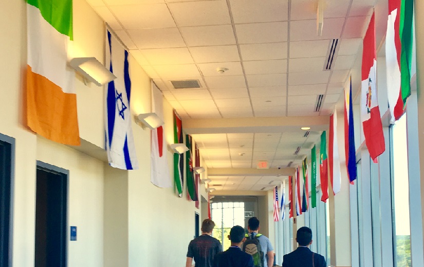 A view of international flags lining a hallway. Four young men walk down the hallway with their backs turned to us.
