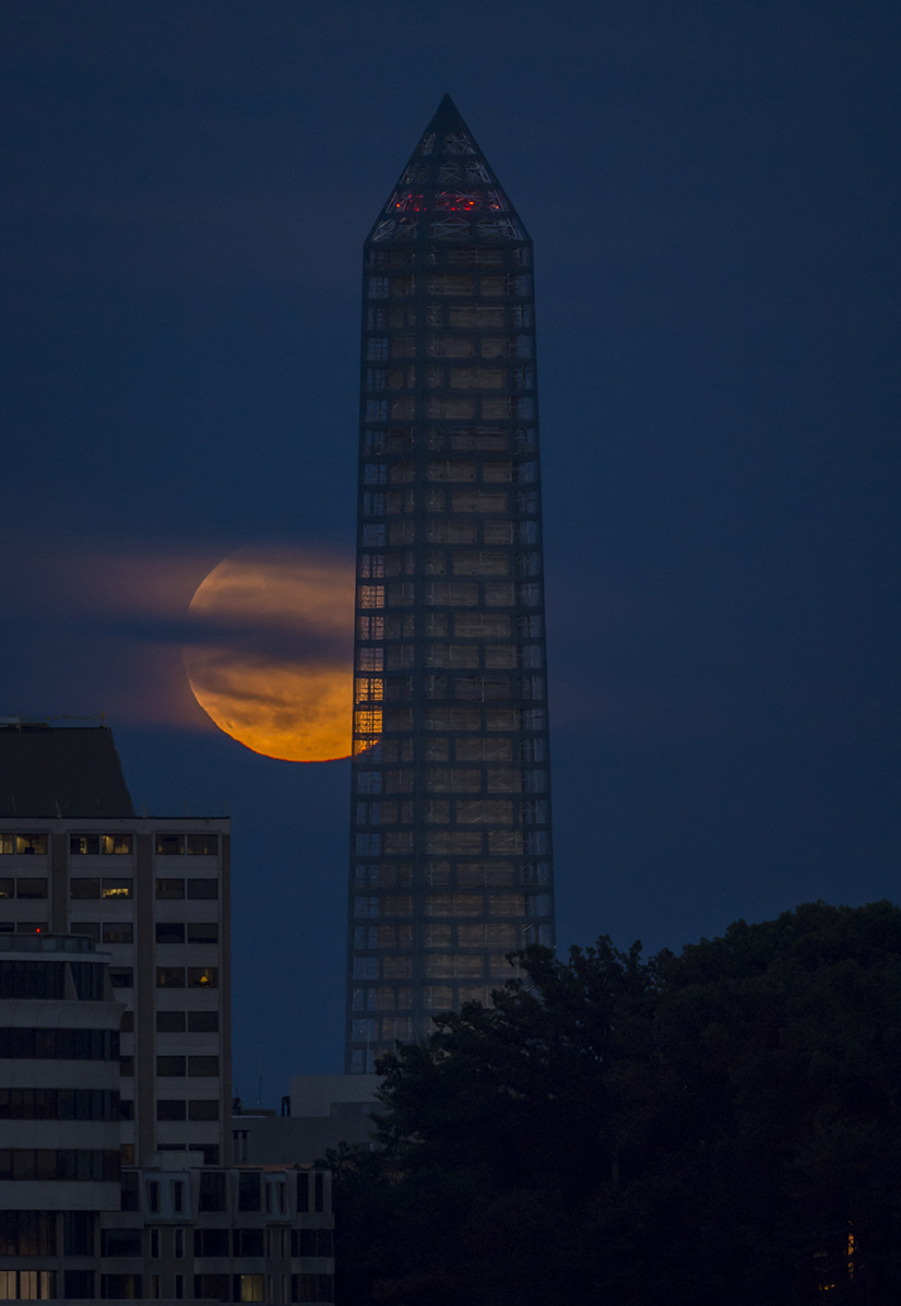 A supermoon rises behind a building with trees in the background.