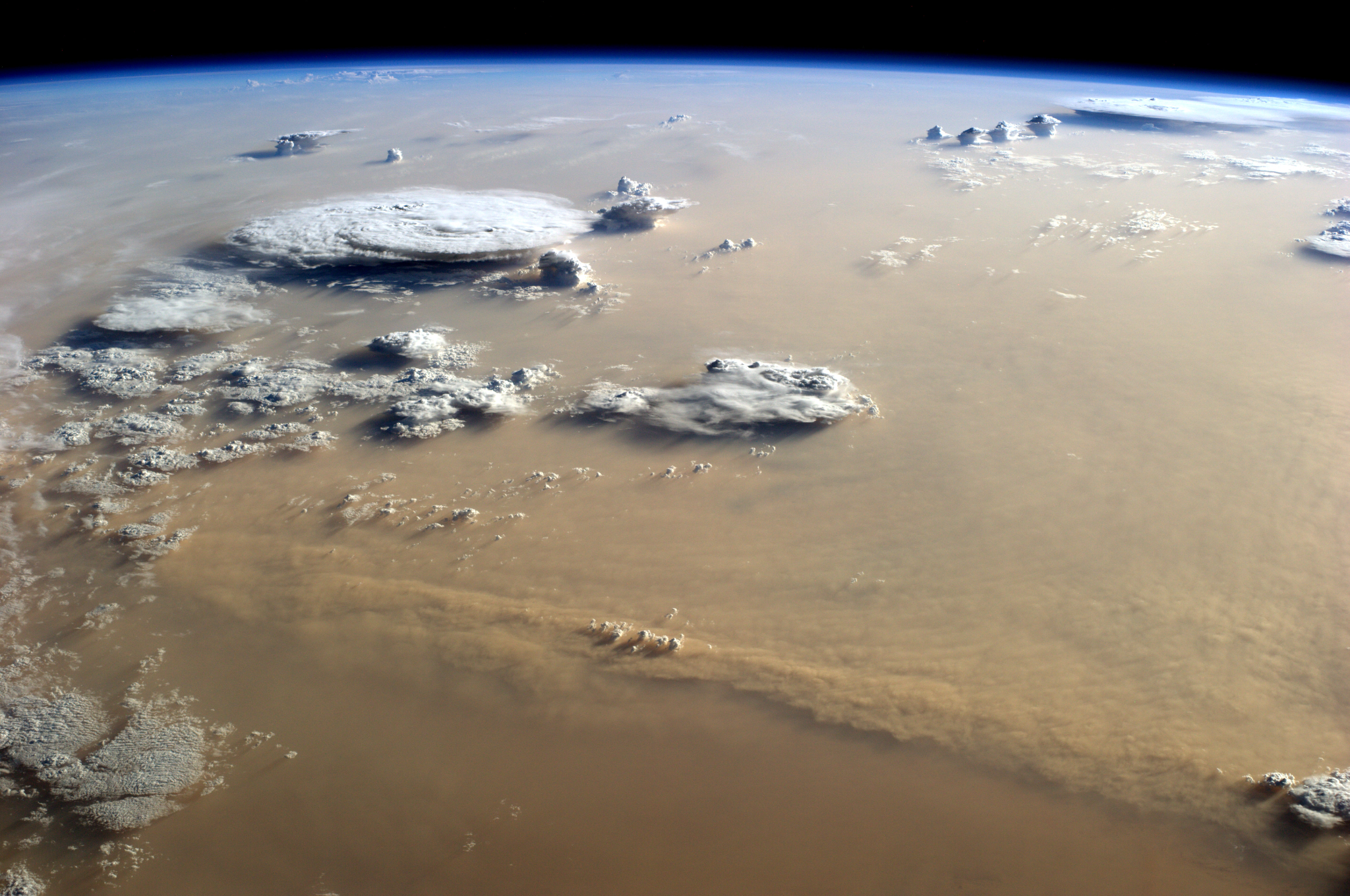 A photograph of Earth from the International Space Station shows a large dust storm coloring the skies brown. Towering thunderstorm clouds rise above the dust. The Earth's blue limb is at the top of the photo. 