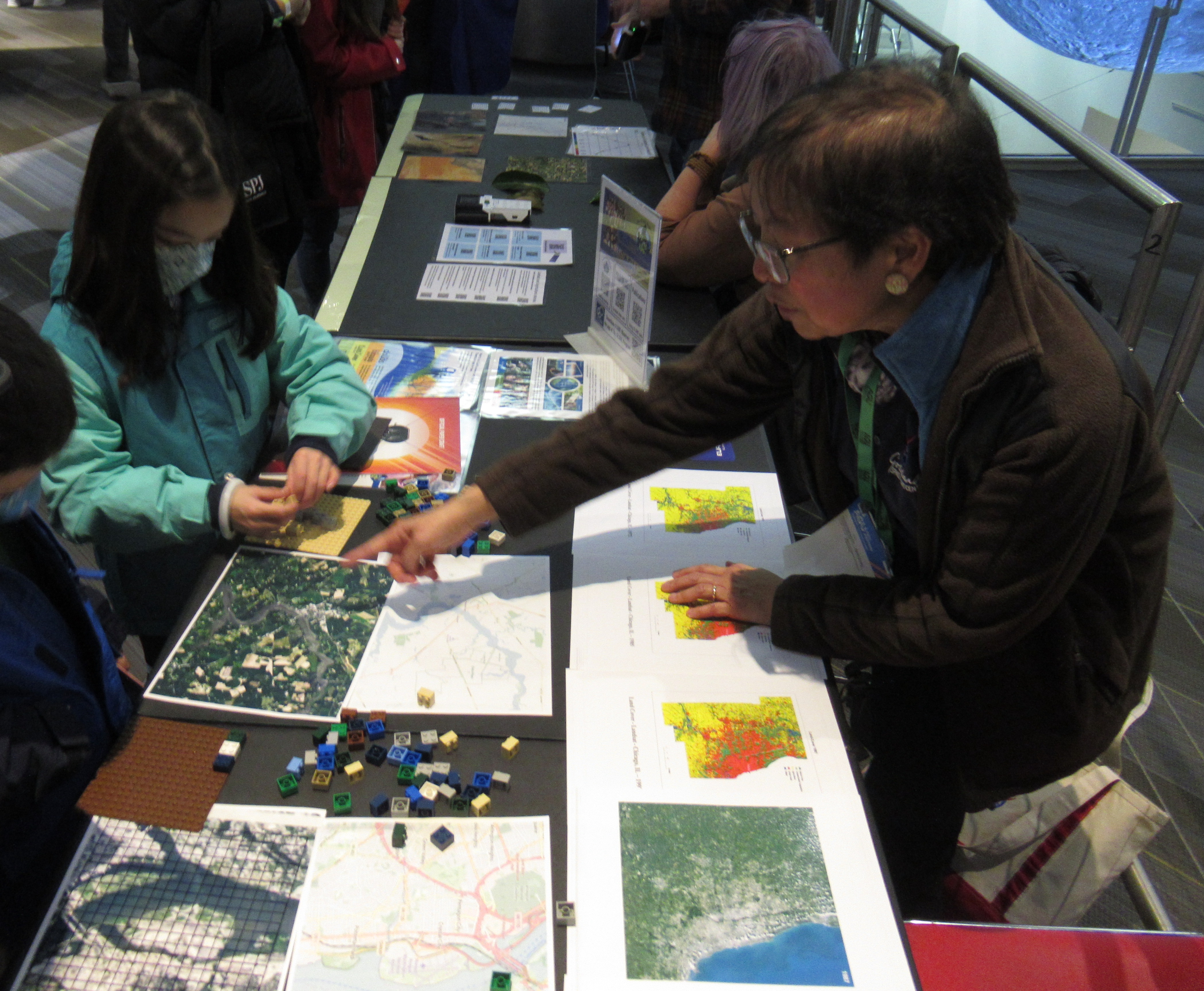 A woman points at a satellite image of the Earth from the Landsat satellite, explaining how the information in the image can be translated into land cover classifications, as demonstrated with LEGO blocks also on the table.