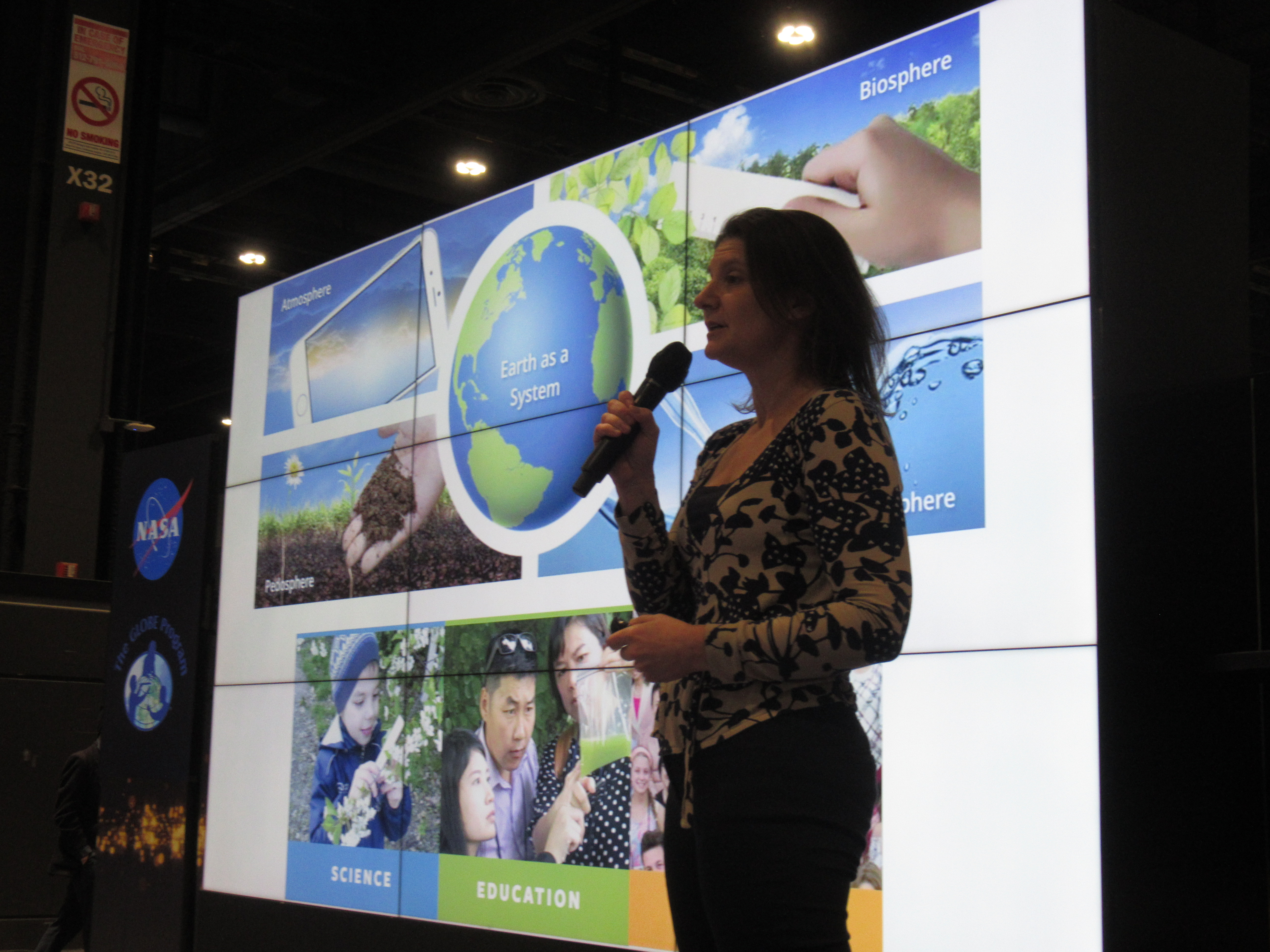 A woman with a microphone presents in front of multiple television screens. On the screen is an Earth as a System diagram with the four spheres of GLOBE protocols, atmosphere, biosphere, pedosphere and hydrosphere.