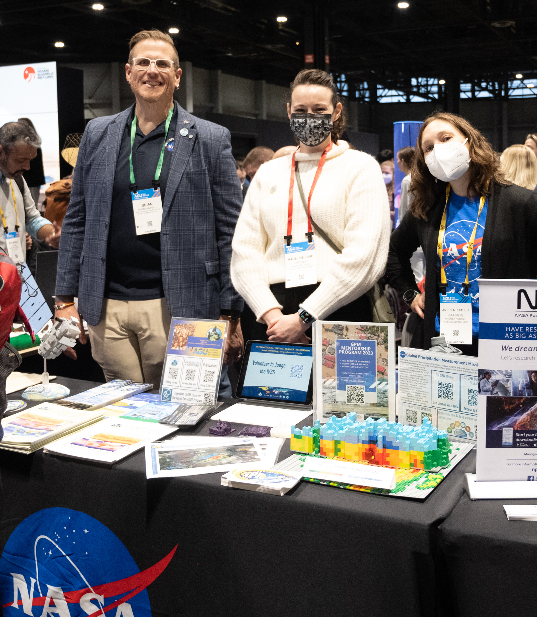 Three people, one man and two women, stand behind a table with a NASA tablecloth. On the table are handouts and resources related to the GLOBE Program as well as materials related to the Global Precipitation Measurement Mission satellite.