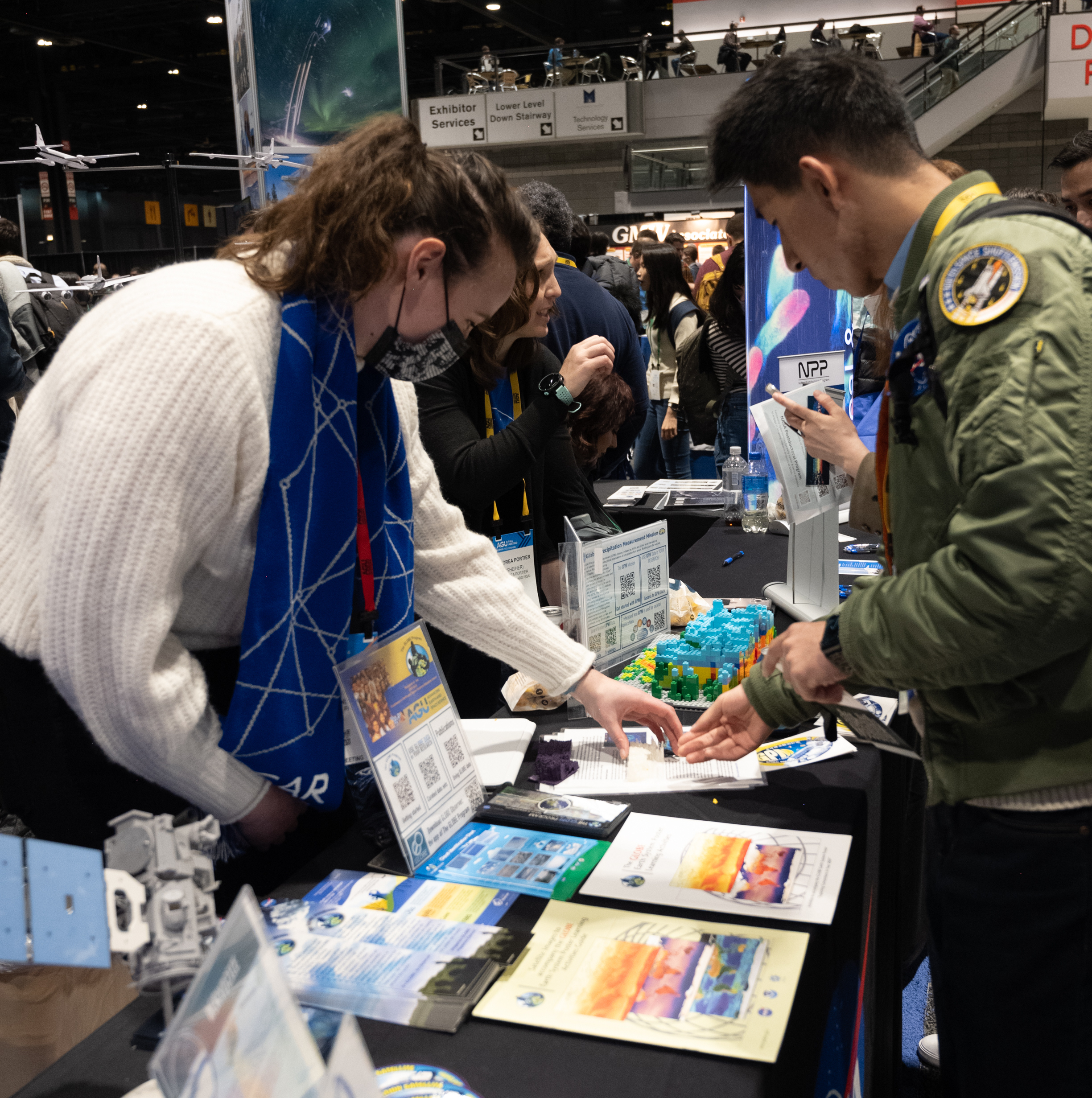 A woman in a white sweater and scarf points at materials on a table while a young man standing on the other side of the table appears to ask questions.