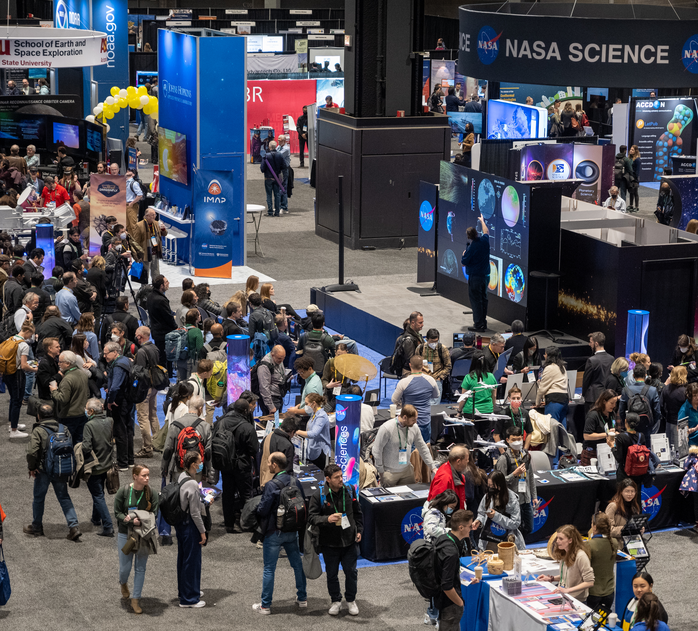 A large group of people crowd around multiple staffed display tables in a large, open room. Behind the tables is an audience sitting facing a set of display screens (the NASA hyperwall) while a presenter gives a talk. Above it all is a hanging sign that says “NASA Science.” Other booths are visible in the foreground and background.