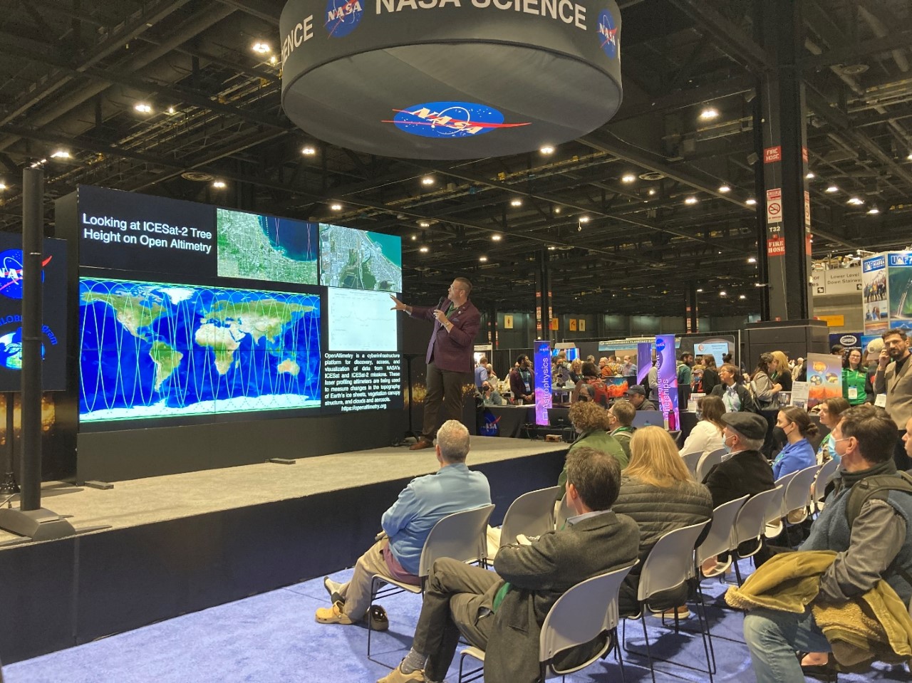 A man points at diagrams on the NASA Hyperwall showing a map from the website OpenAltimetry of the satellite swath locations for ICESat-2 data related to tree height, as well as specific examples of zooming in to get data from a particular place.