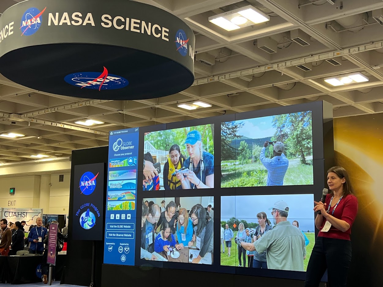 A woman stands in front of a large area of display screens. The left side shows the NASA and GLOBE Program logo. In the middle is the main landing screen of the GLOBE Observer app. To the right are four different pictures of people collecting various types of environmental science data with GLOBE. Above it all is a large circular sign that says "NASA Science."