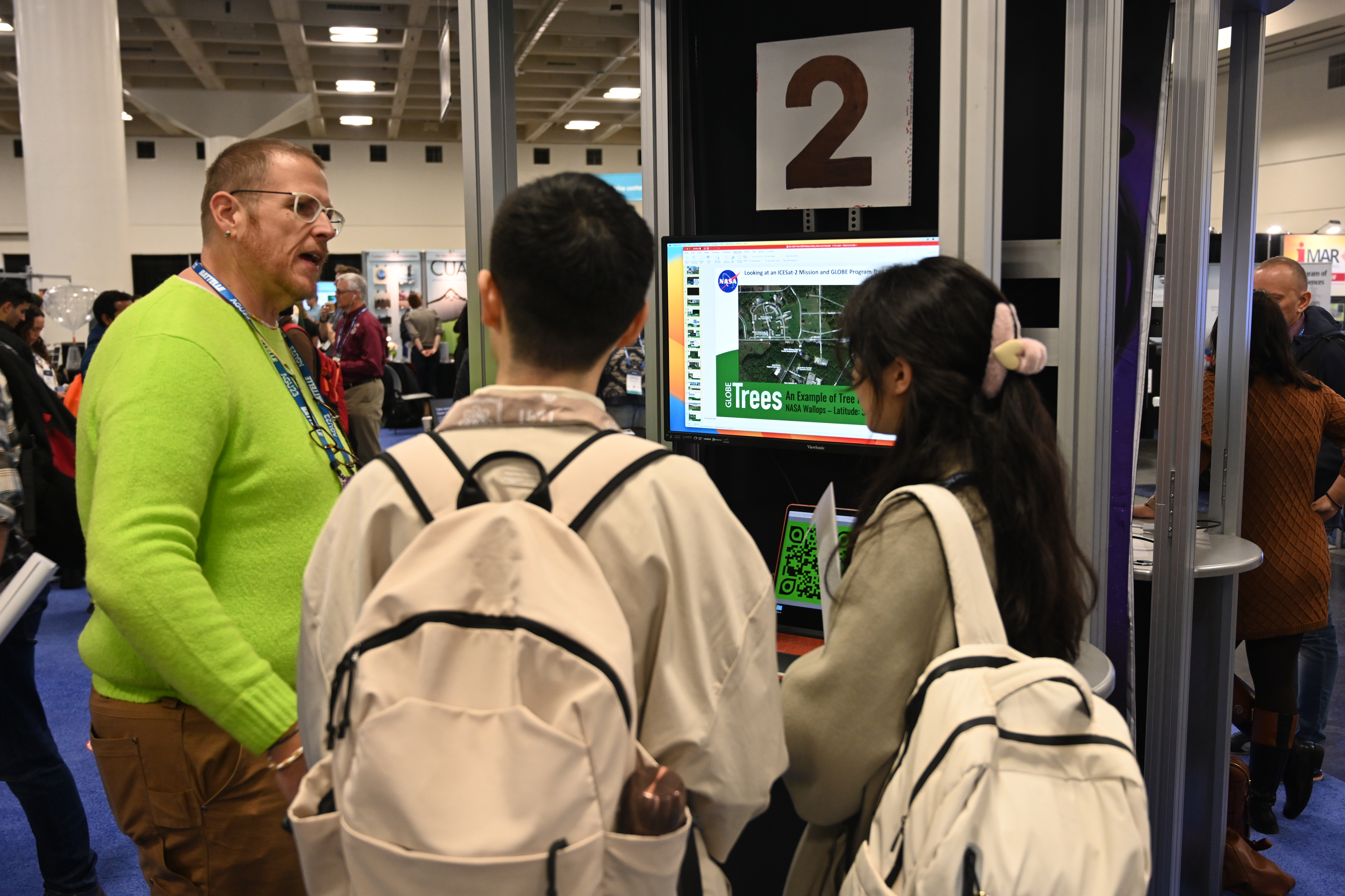 A tall man in a bright green shirt stands next to a monitor in a demo kiosk that displays information from the GLOBE Trees tool. He's talking to two visitors wearing white backpacks and listening attentively.