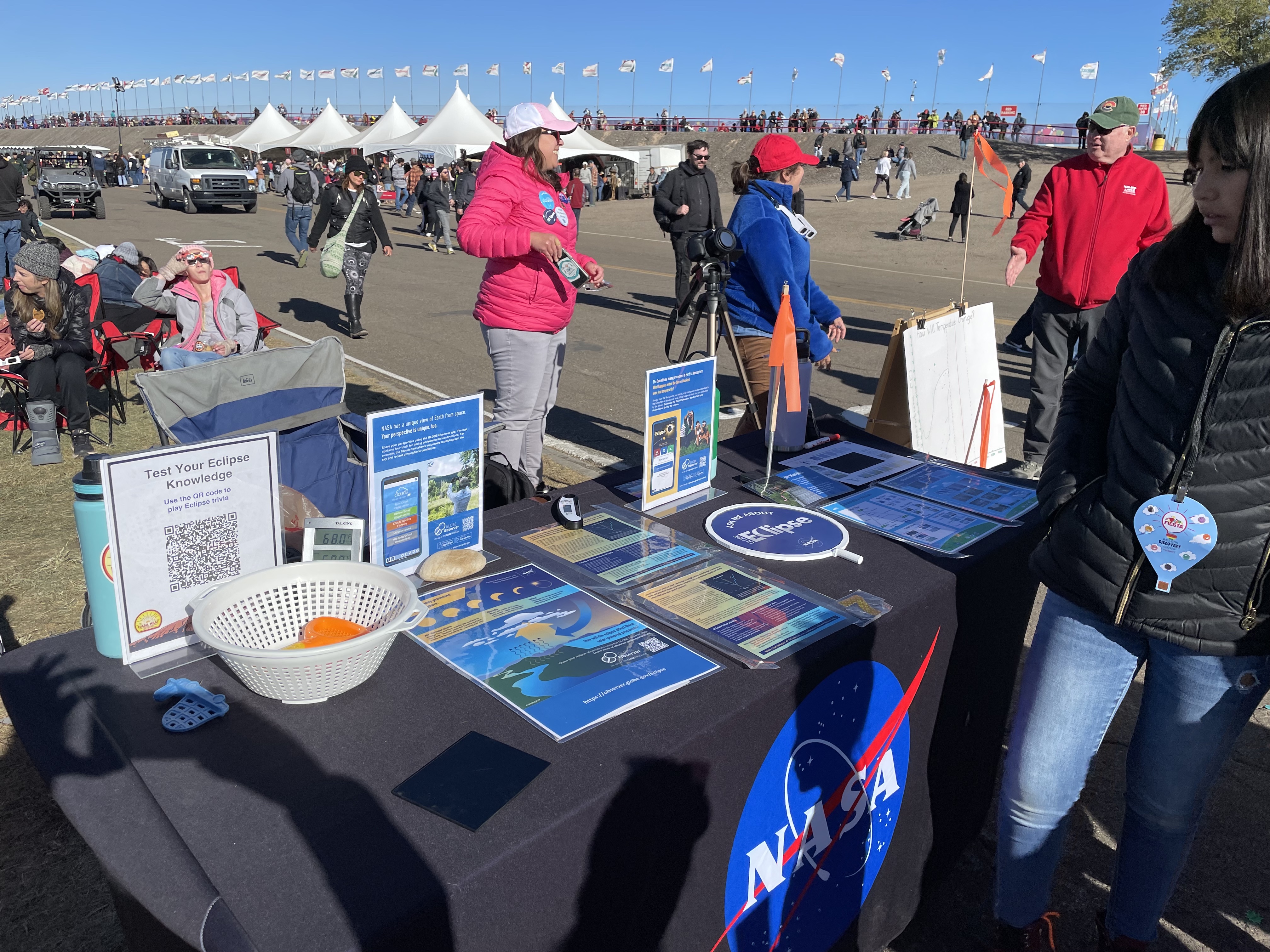 A tabletop display includes a colander and toys that have holes that cast shadows in the shape of the eclipse. The table also includes signs about GLOBE eclipse and clouds. Text on the signs is not legible. A woman is looking at the table while people in the background are watching the eclipse through solar viewing glasses. 