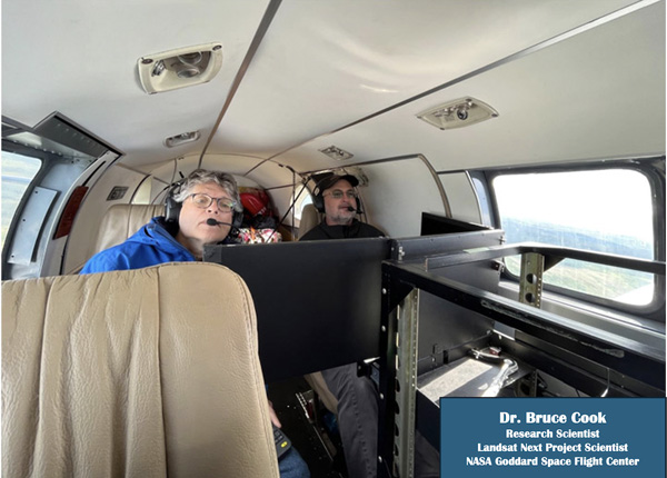Two men with glasses and headphones ride in the back of a small plane while monitoring an array of computer displays.