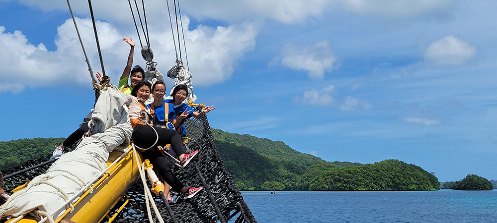 Four teens sit in the bow net of a large sailing ship. In the background is a green island. 