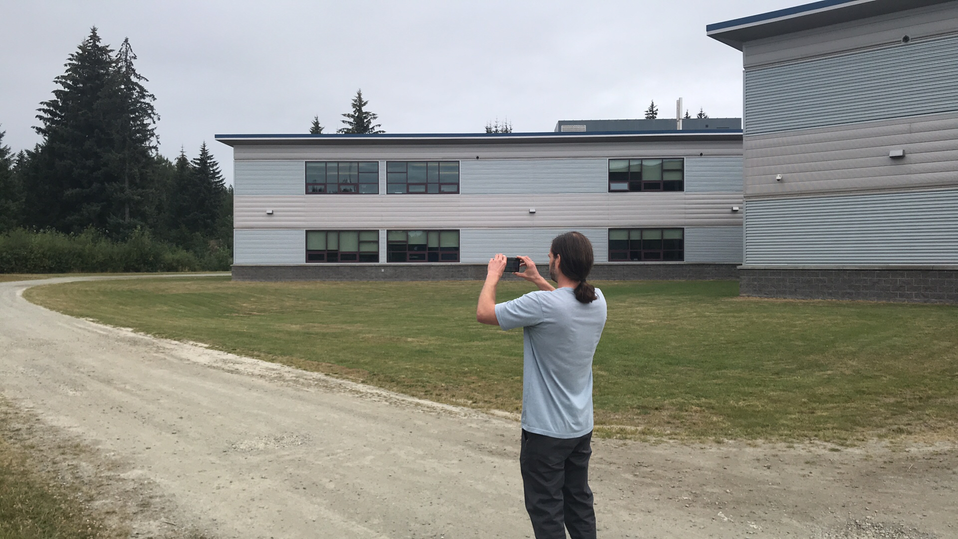 A man holds a phone horizontally and is photographing a dirt road, building, grass and the edge of an evergreen forest.