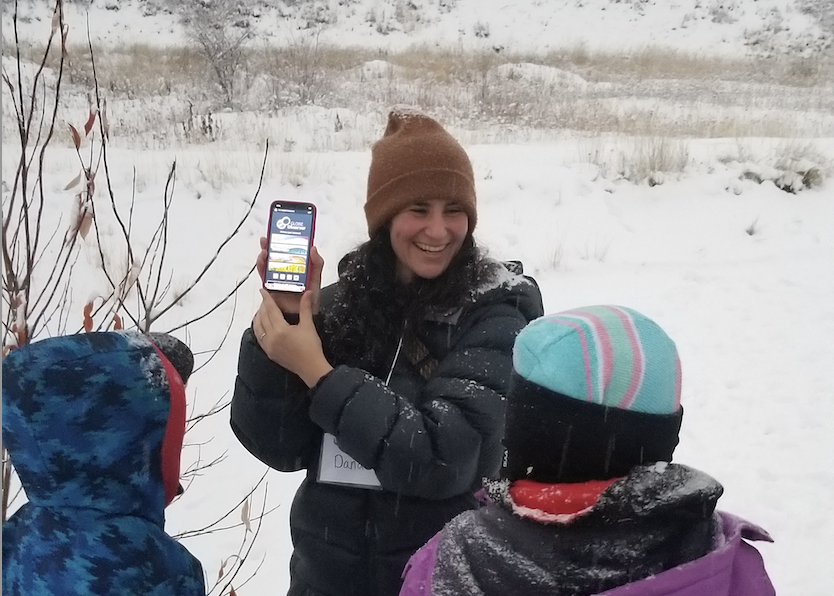 A photo of a woman in a winter coat and hat showing a phone screen to two children. The screen shows the GLOBE Observer app. All are standing outside on a snow-covered landscape. 