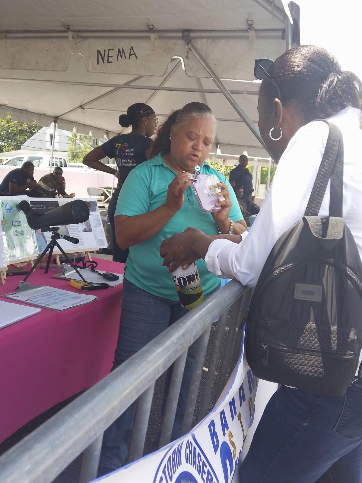 Monique holds a phone with a clip on microscope over the camera lens. She is demonstrating how to use the microscope lens to photograph mosquito larvae to a woman attending an outdoor event. 