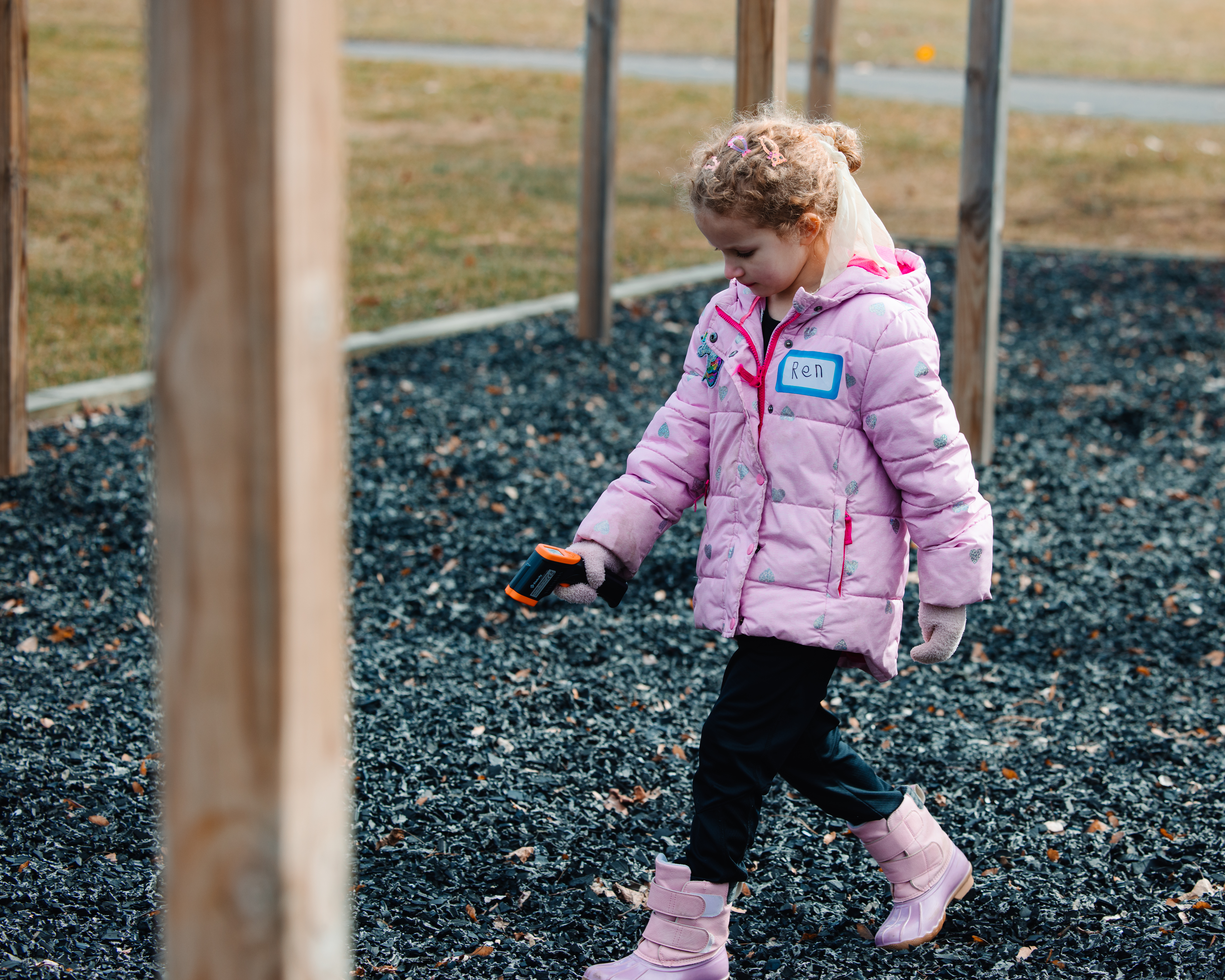 An elementary-aged girl is walking across a contained area covered in bark or some other dark ground cover. The contained area is surrounded by grass. She holds an infrared thermometer pointed at the ground to measure surface temperature.