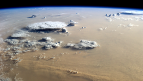 A photograph of Earth from the International Space Station shows a large dust storm coloring the skies brown. Towering thunderstorm clouds rise above the dust. The Earth