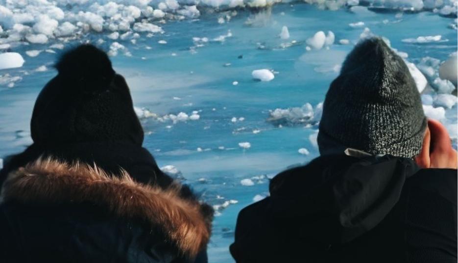 Two people in winter clothing look at ice on a river or lake.