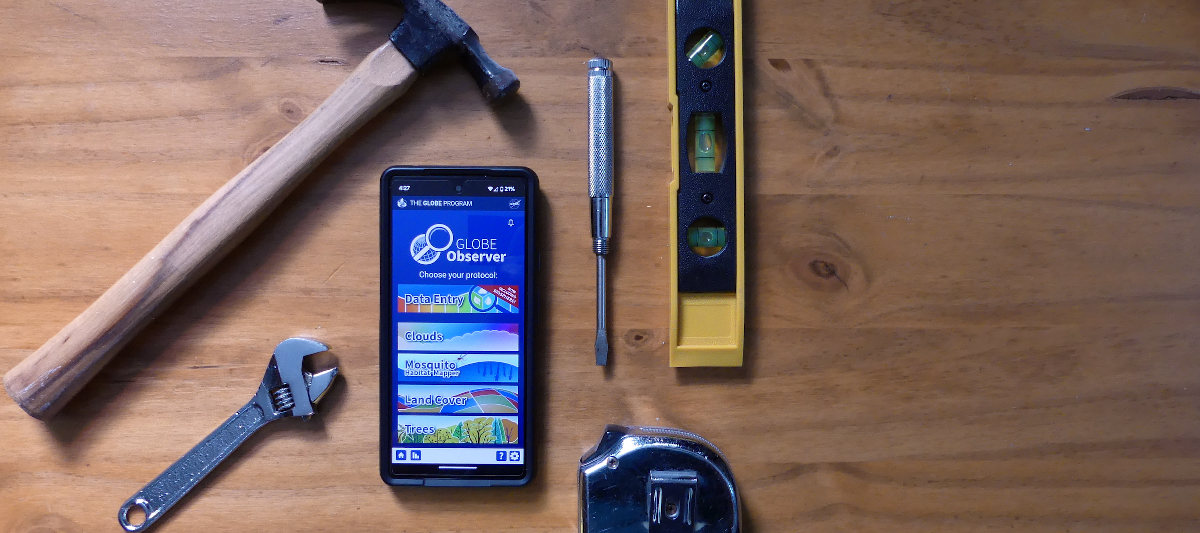 Various tools, a hammer, wrench, screwdriver, tape measure, and level, surrounding a phone with the screen showing the GLOBE Observer app.