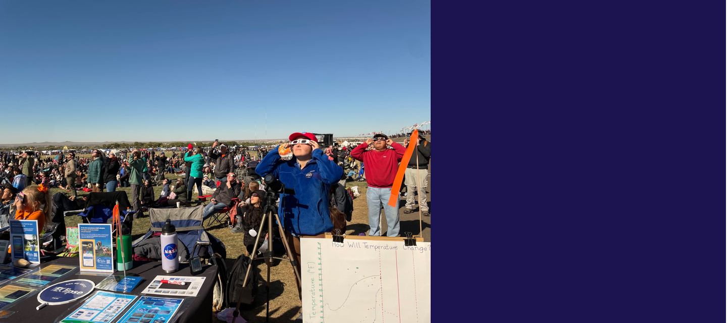 A table with materials about GLOBE Eclipse and a part of a paper chart with temperature data on it, in front of a large crowd. People are sitting and standing, and many are holding solar viewing glasses up to their face, looking at the Sun. The sky is clear of clouds.