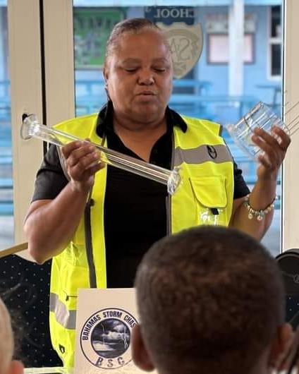 A woman wearing a yellow safety vest is holding a rain gauge while speaking to a group of children. A sign that says "Bahamas Storm Chasers" is in front of her.