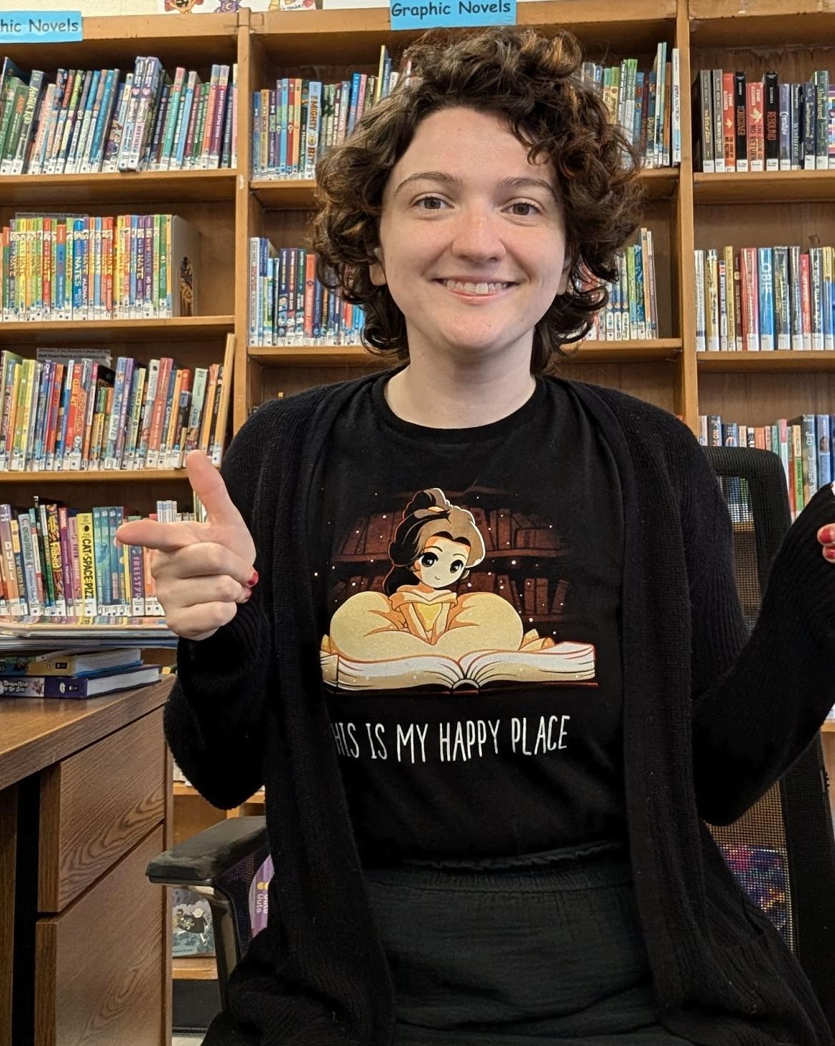 A young White woman with brown eyes and short, curly brown hair sits in front of bookcases, smiling. Her t-shirt has a manga-style character in a ball dress reading a book, and the text 