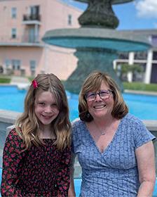 A girl and her grandma sitting next to each other in front of a fountain, smiling. It's a sunny day with some clouds.