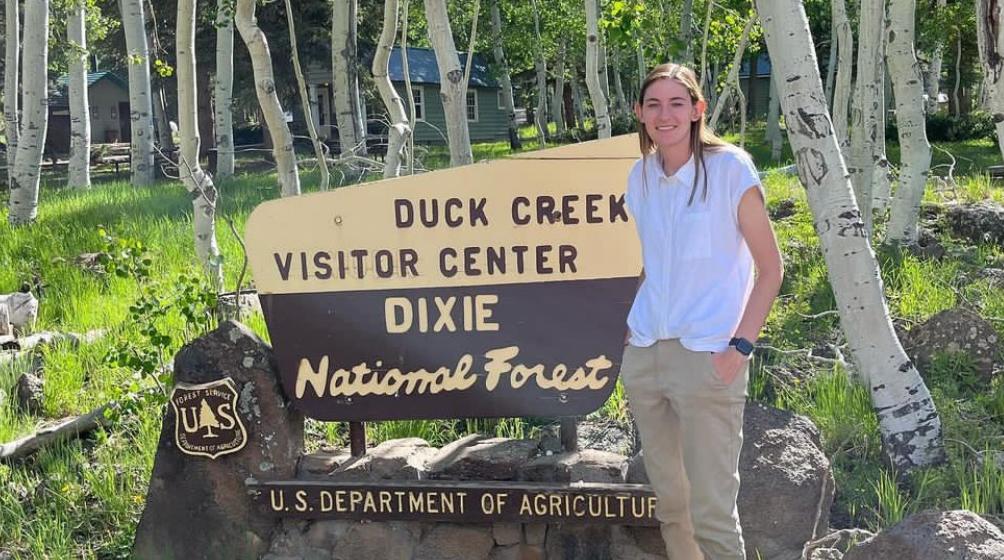 A young woman stands next to a sign that reads “Duck Creek Visitor Center Dixie National Forest U.S. Department of Agriculture.” An aspen tree forest surrounds her.