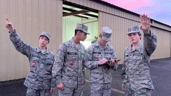 Four youth in Civil Air Patrol uniforms collecting data. Two are pointing at the sky, and a third is making notes on a clipboard, while a fourth looks on. In the background is an aircraft hanger.