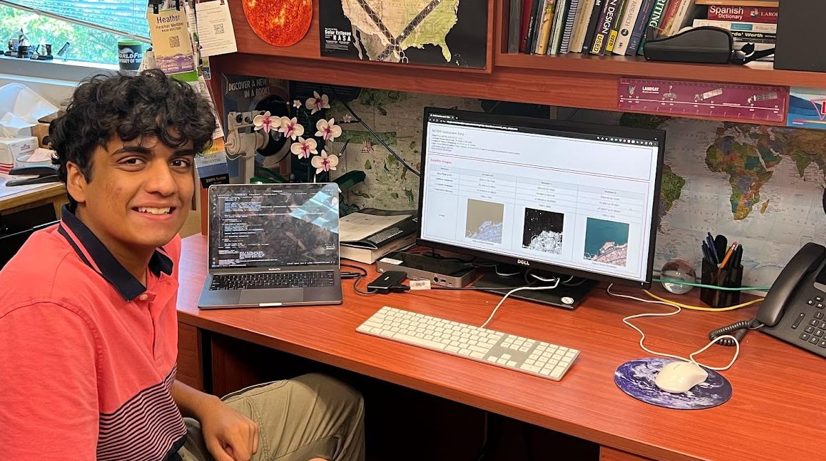 Intern Sidd Jasti sitting at a desk with computer screens showing his code and the satellite comparison table he worked on for his project.