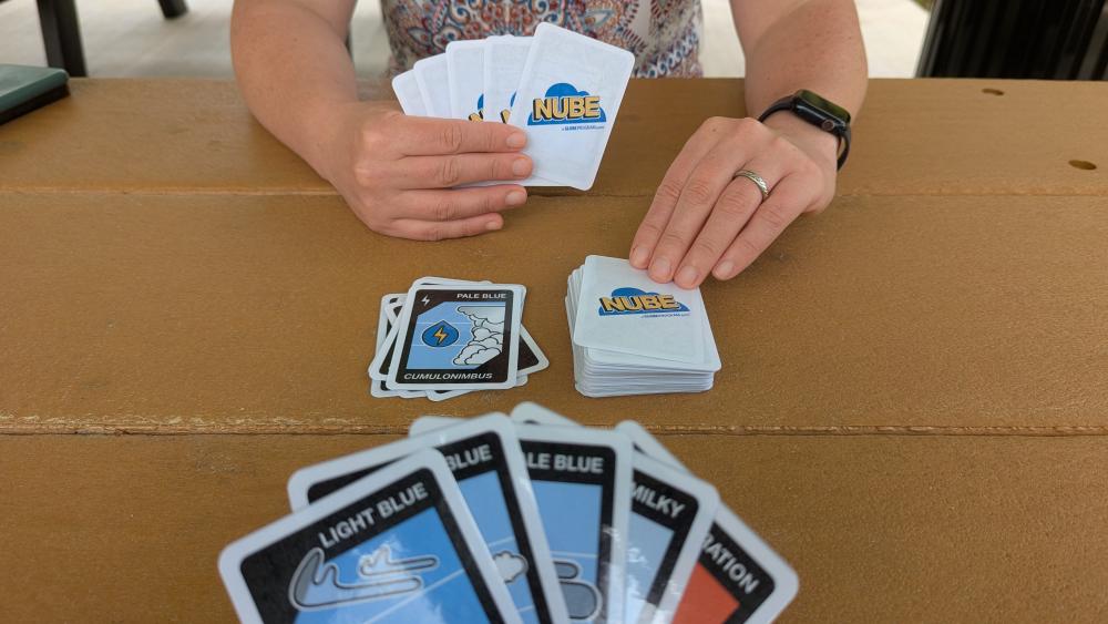 A table showing a card game going on - one person's holds their cards in one hand, and are about to draw from the deck, the other person's hand is visible in the foreground.
