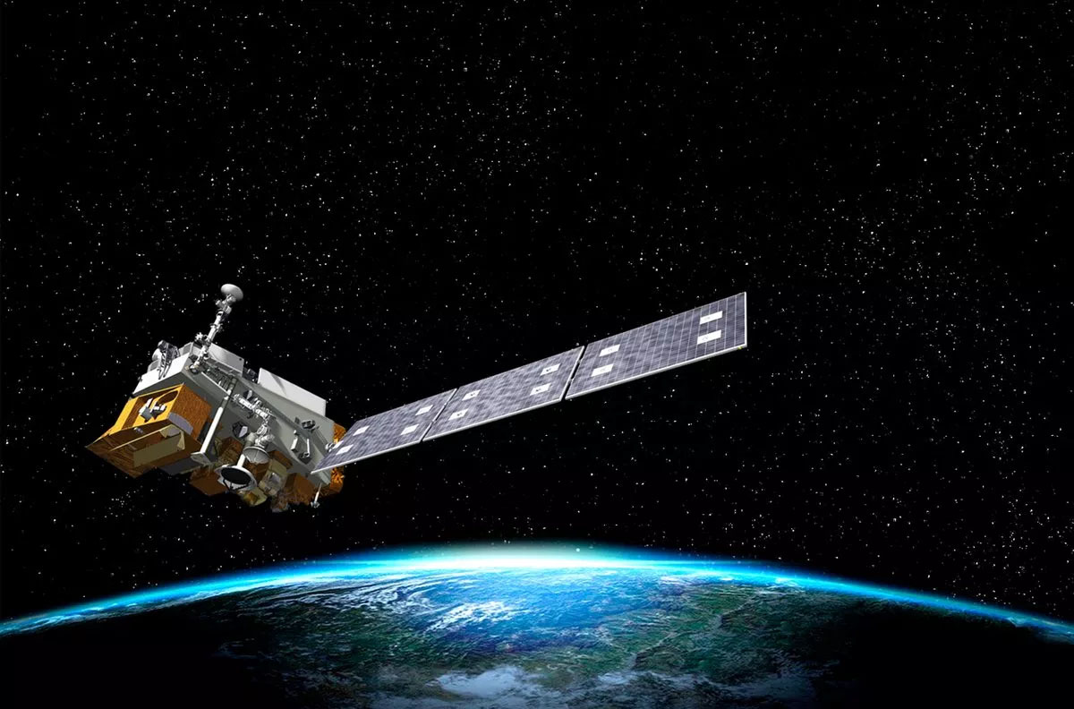 A satellite in orbit over the Earth. The roughly cubic main part of the satellite is to the left, with protrusions of various instruments protruding from it. To the right, a three-section solar panel array extends. In the upper background is a the darkness of space with stars, above a hemisphere of Earth showing land, water and the thin sliver of atmosphere around the planet.