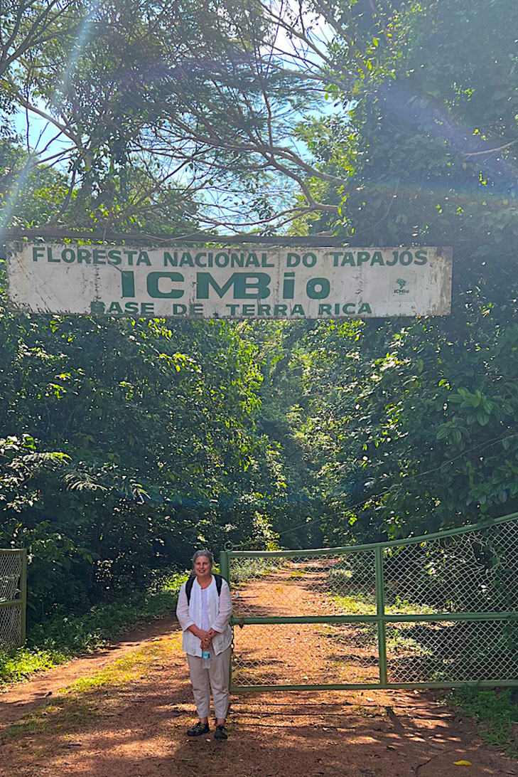 A woman standing in front of a gate on a road leading into trees. A sign above her head says, "Floresta Nacional do Tapajos, ICMBio, Base de Terra Rica."