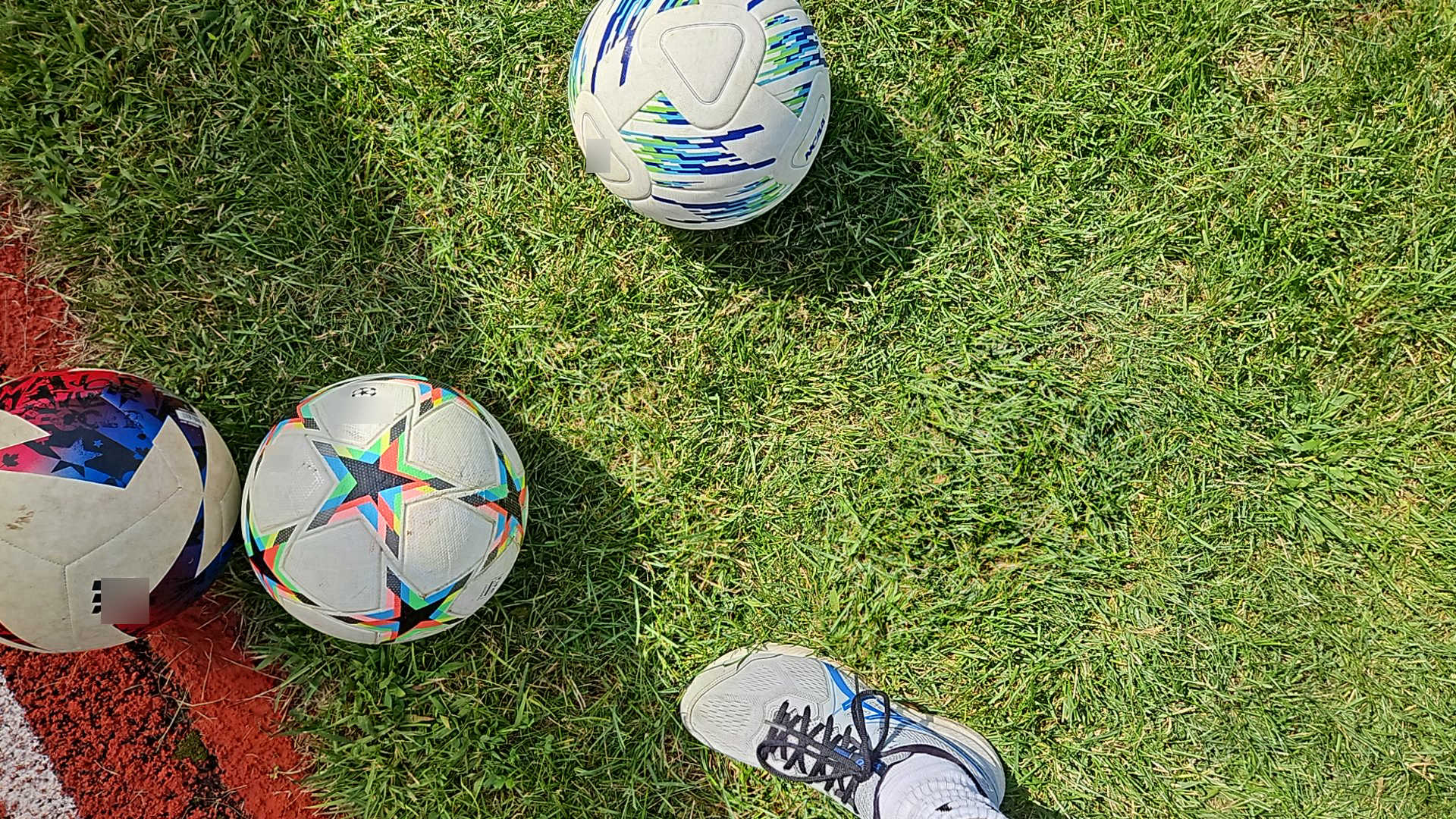 Three soccer balls on grass and a foot in a tennis shoe.