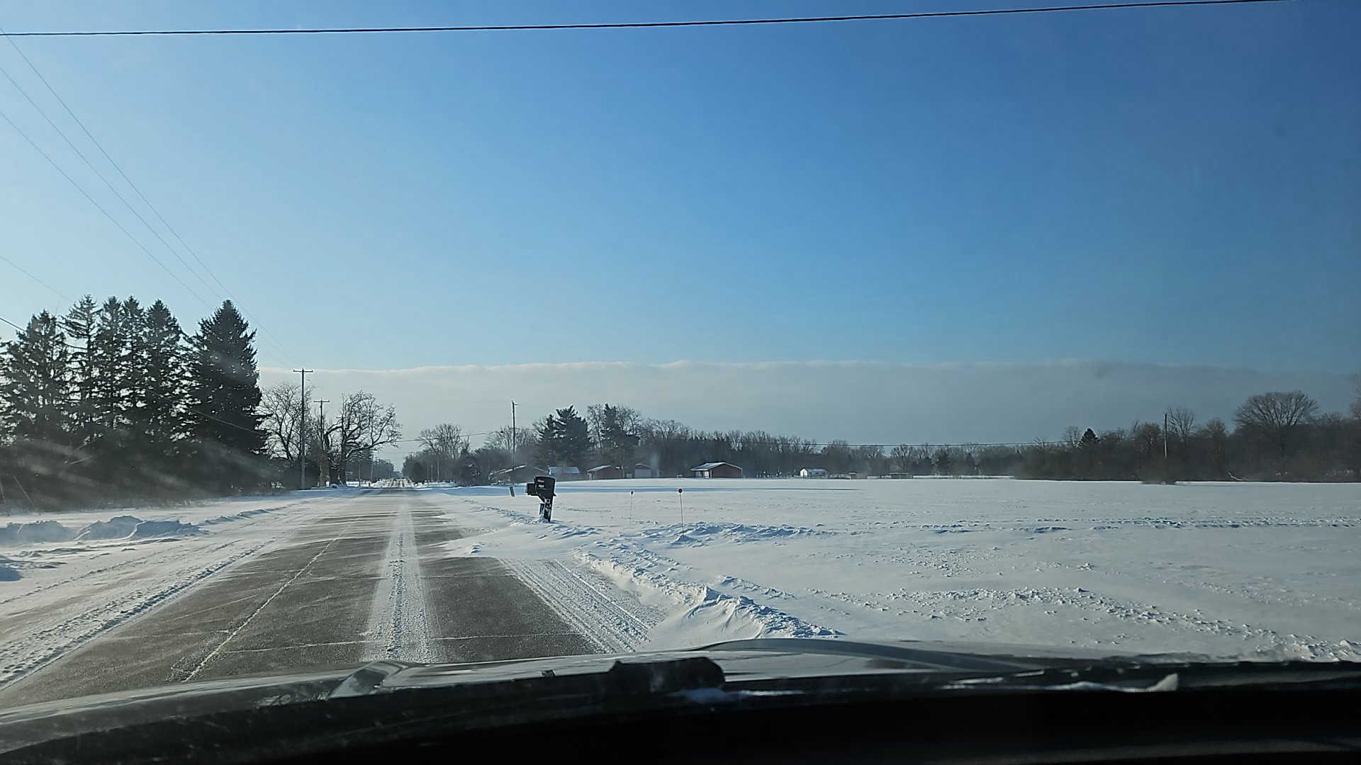 Landscape scene showing snow drifting across a roadway to the left of a snow-covered field.
