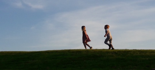 Two children walking on grass are silhouetted against a blue, slightly cloudy sky. 