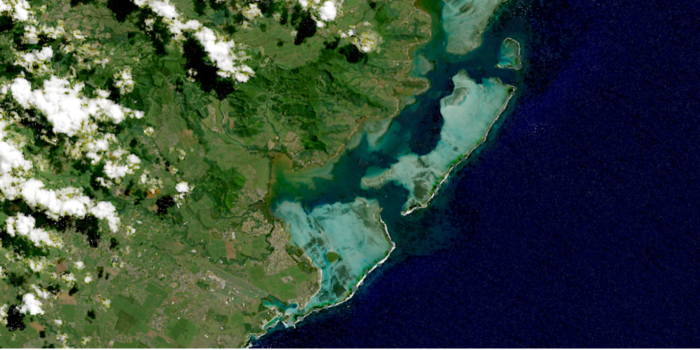 Satellite image of a coastline showing green vegetation partially covered by clouds and a deep blue ocean. Where the land and sea meet, there is bright turquoise water dotted with vegetation and outlines by barrier islands.