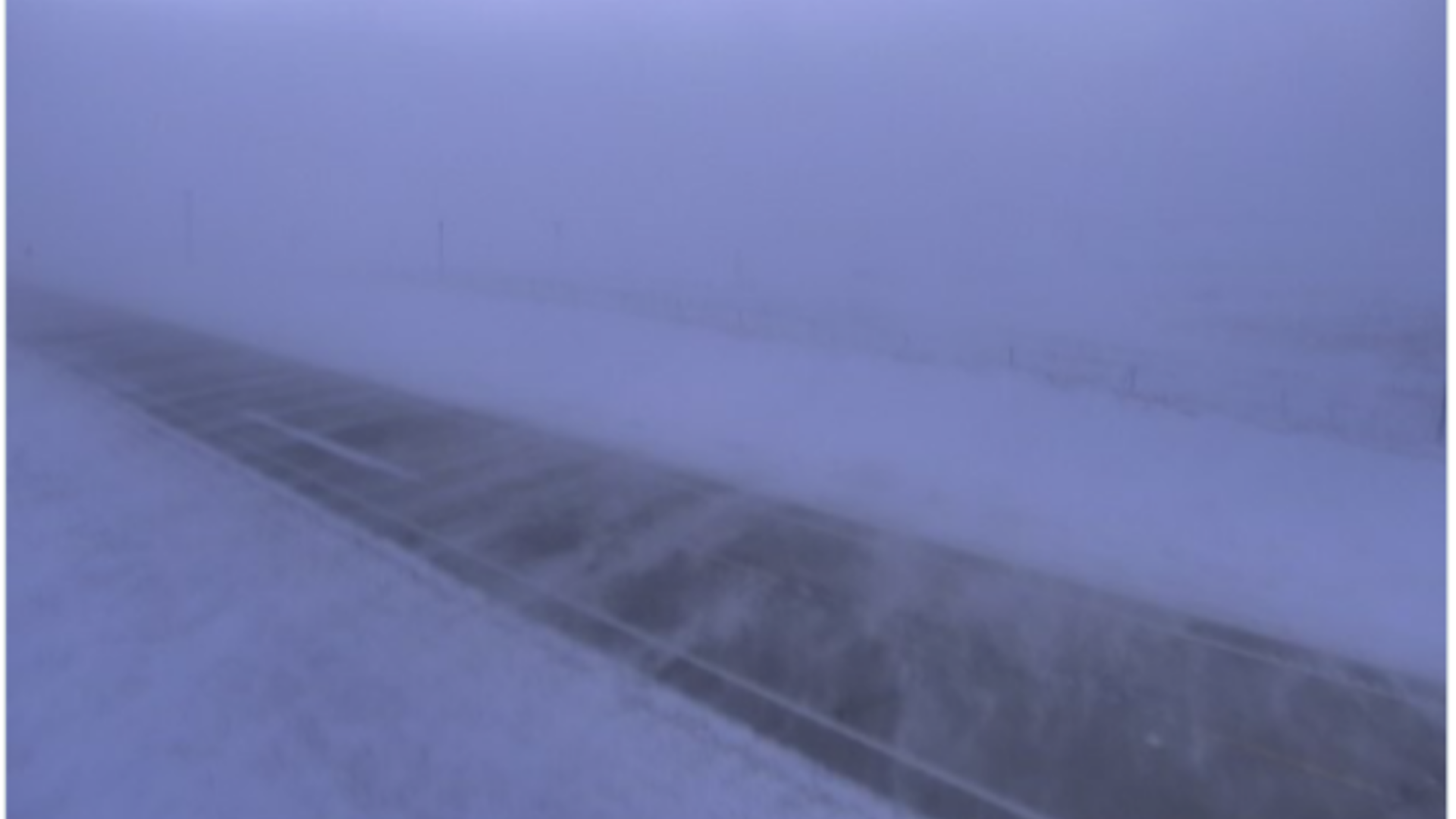 A snow-covered landscape is blurred by blowing snow. A narrow road is in the center of the image.