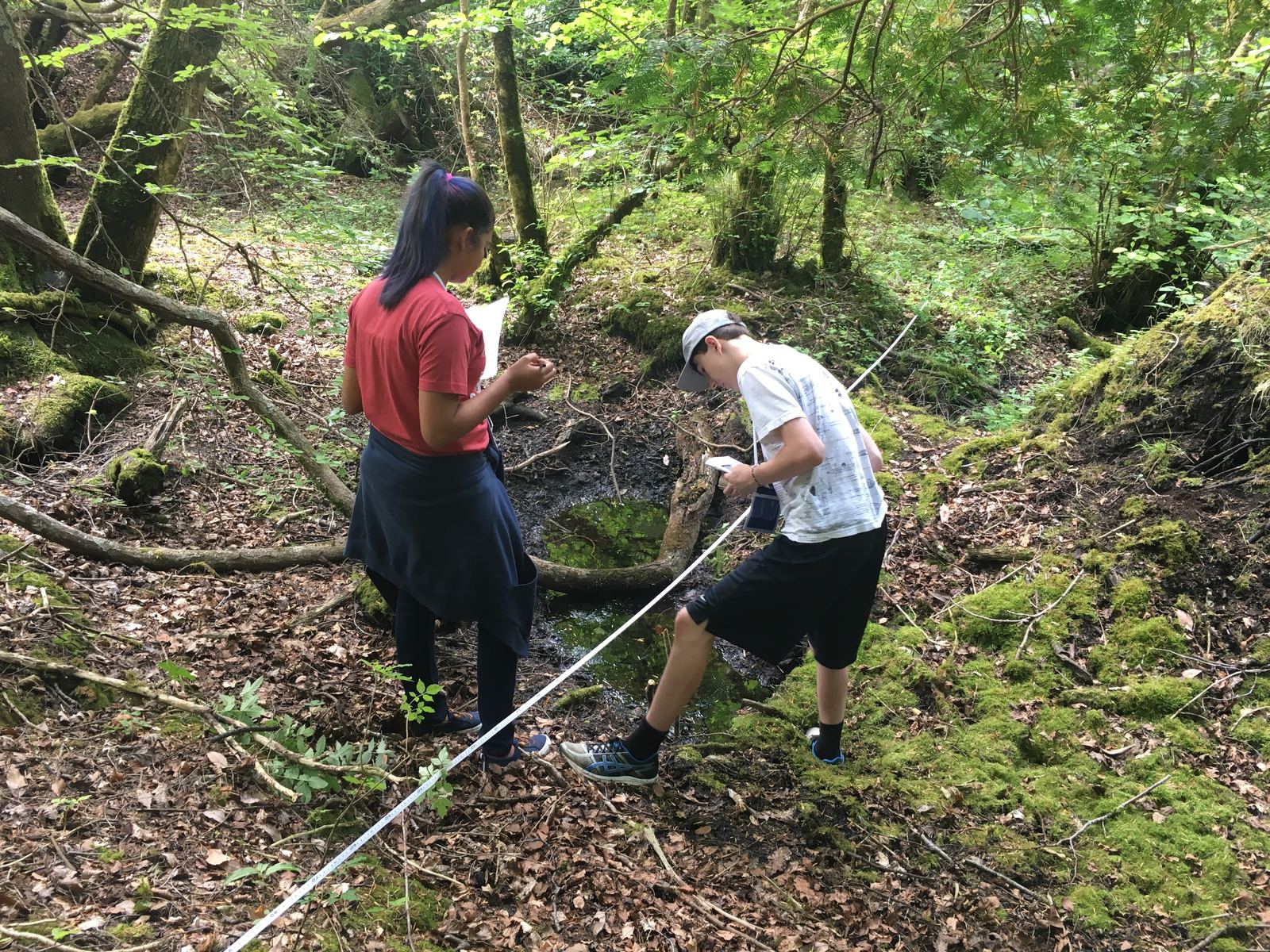 Two teens stand in a forest looking at a long tape measure. Both hold paper.